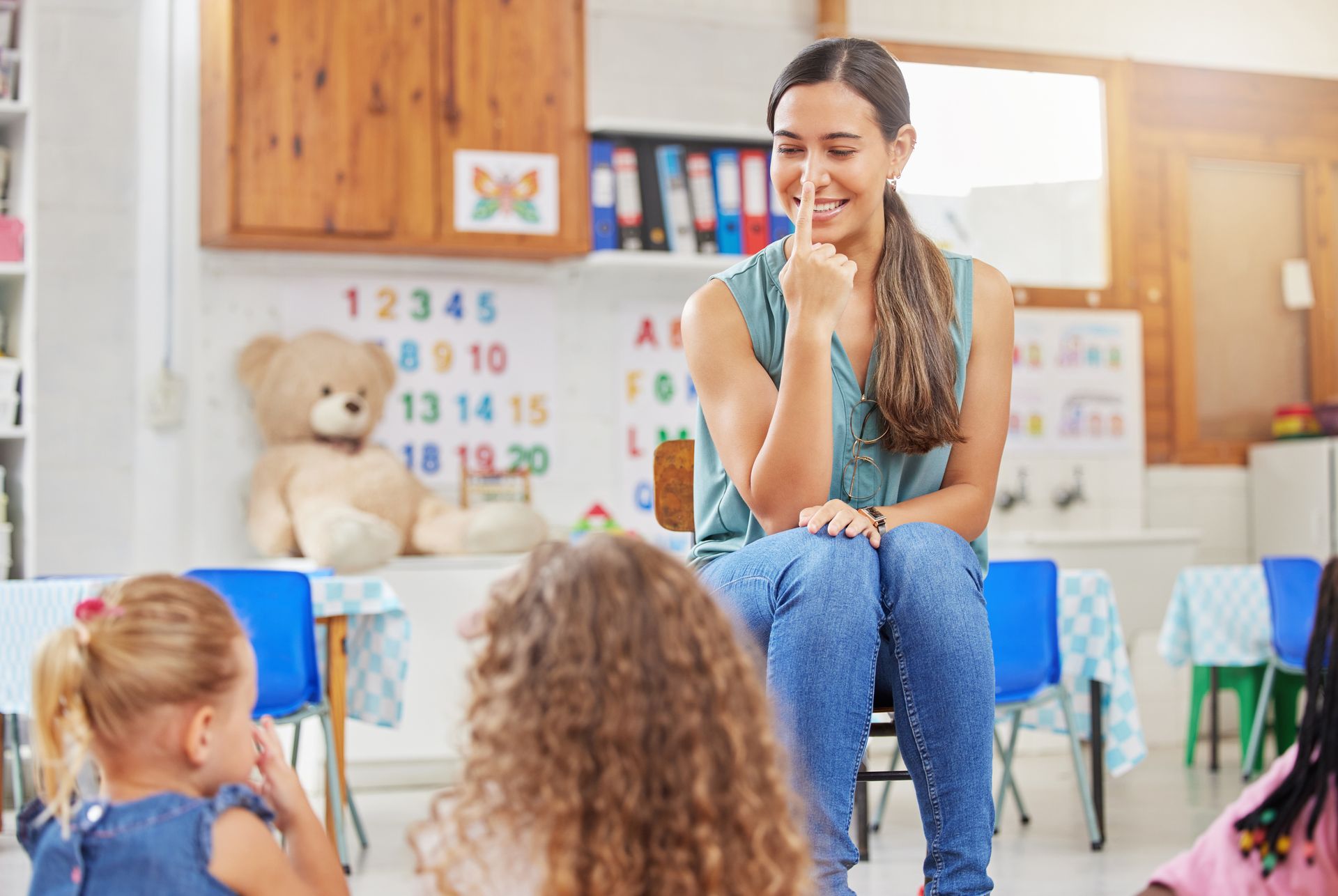 Teacher in a classroom, with hand on chin, smiling at children seated in a circle.
