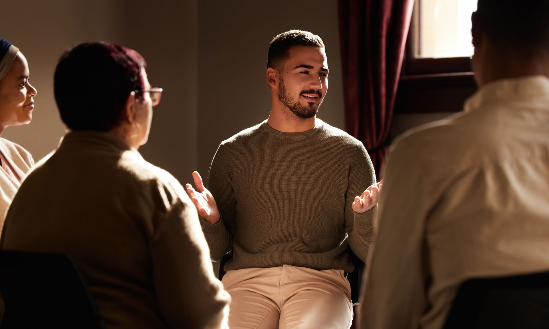 Group therapy session: Man speaking with hands raised, sitting in a circle with three others in a softly lit room.
