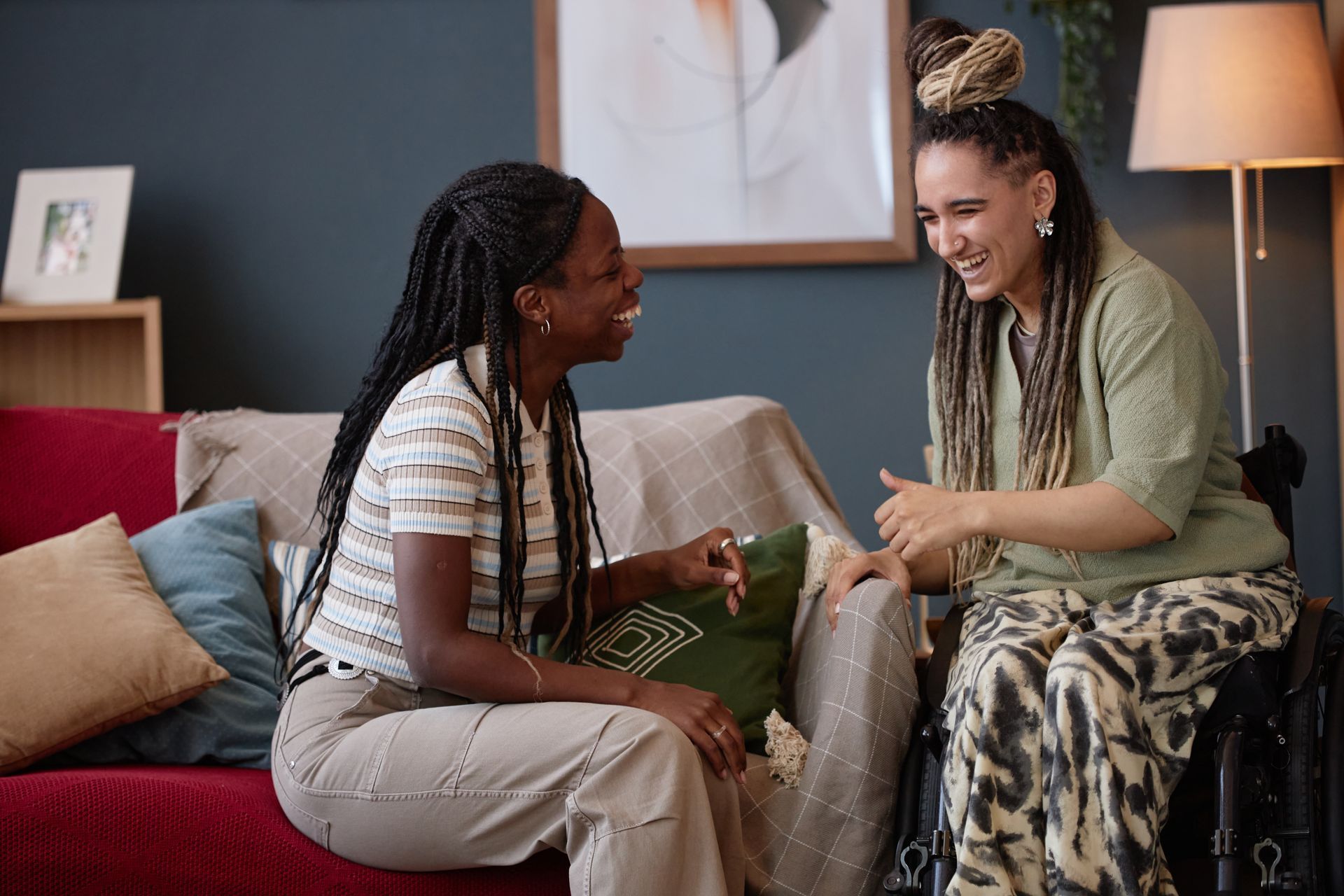 Two women laughing on a red couch, one in a wheelchair, indoors.