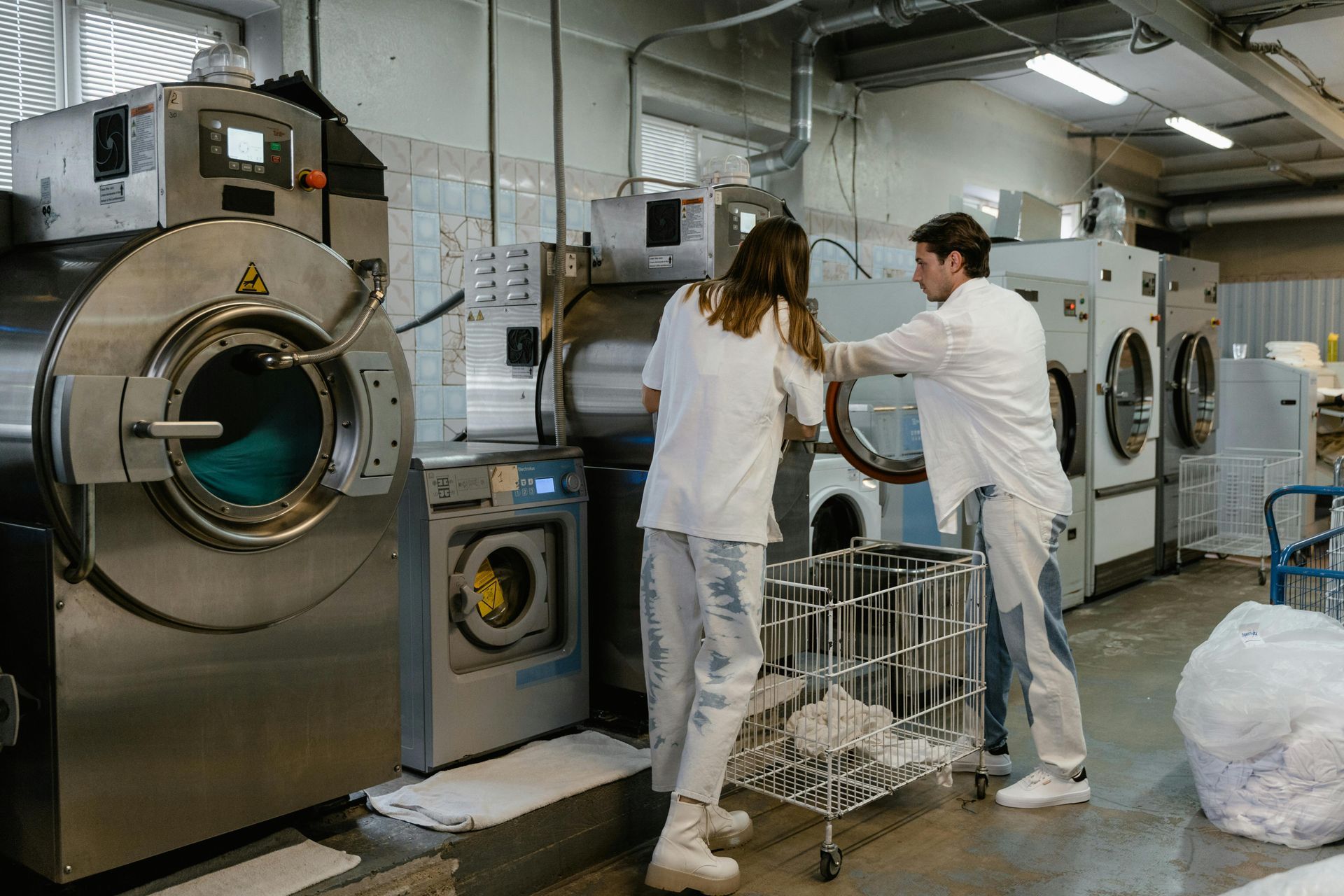 Two people in a laundry facility working with industrial washing machines, placing items in a cart.