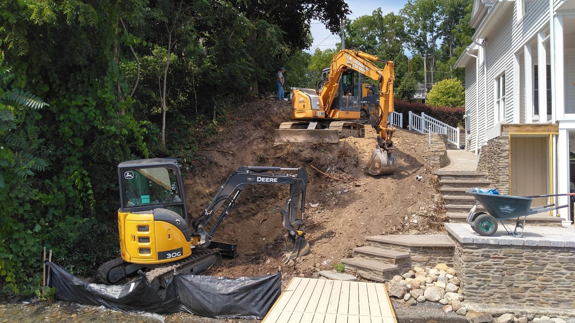 Two excavators are working on a hill in front of a house