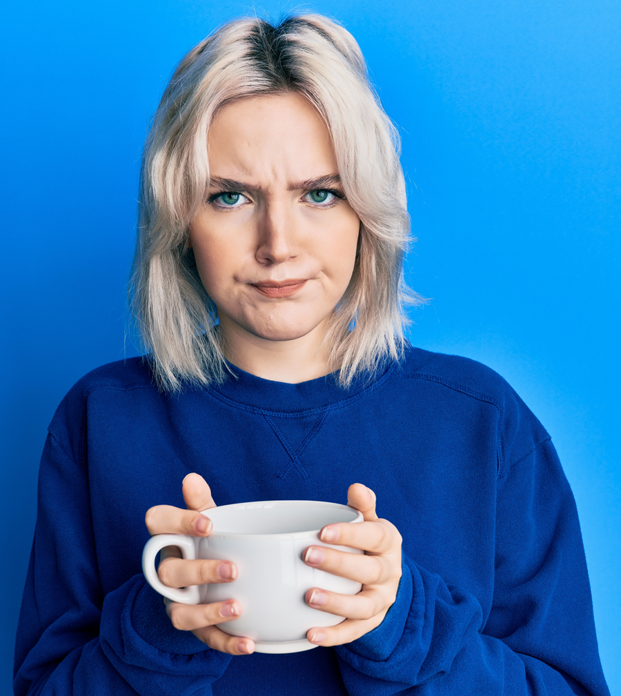 Blonde woman in blue sweater holding white mug, frowning against a blue background.
