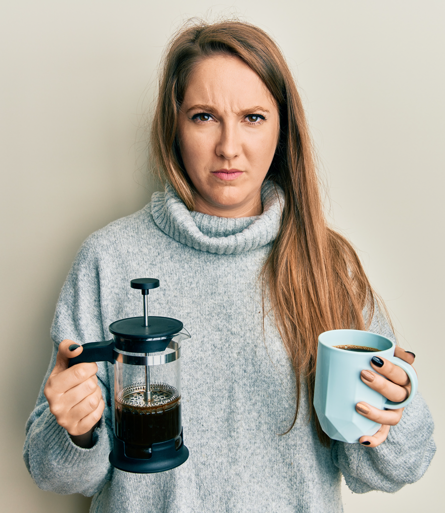 Woman with long blonde hair frowns, holding coffee press and mug.