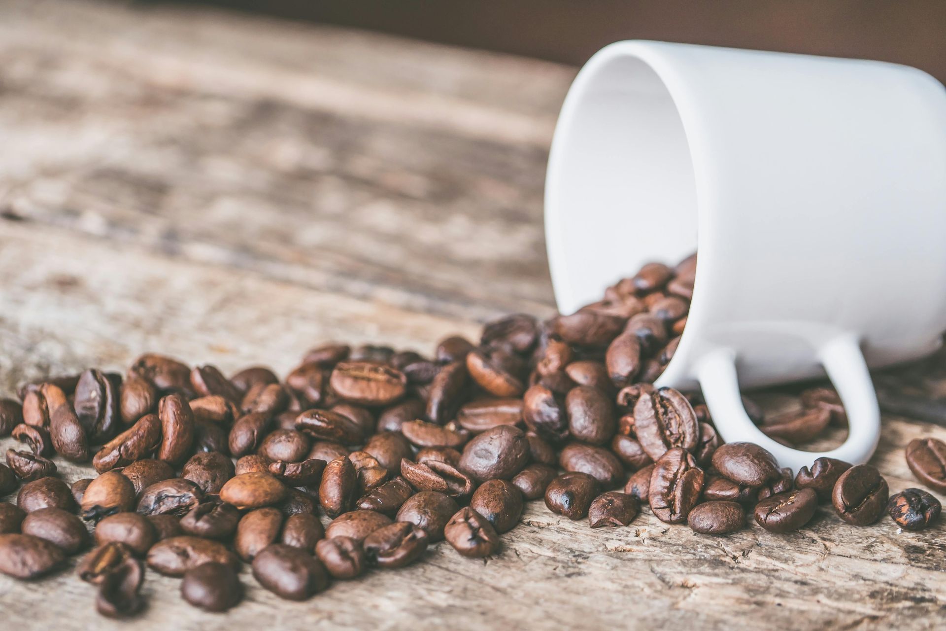 Coffee beans spilling from a white mug on a wooden surface.