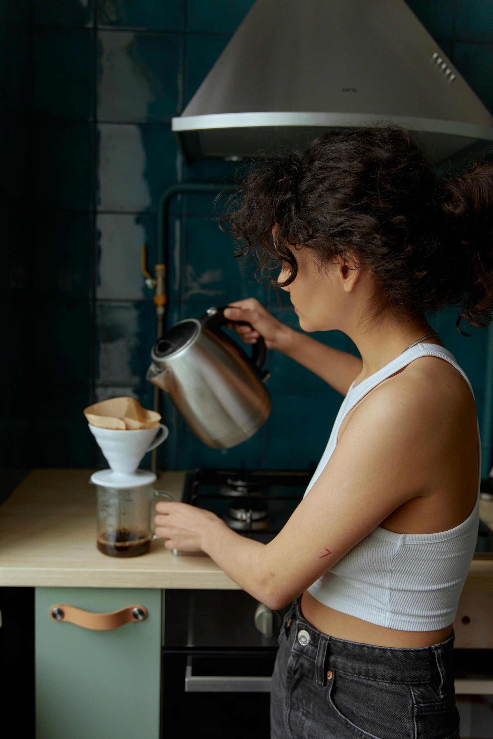 Woman pouring hot water from a kettle into a coffee filter. Kitchen setting with teal backsplash.