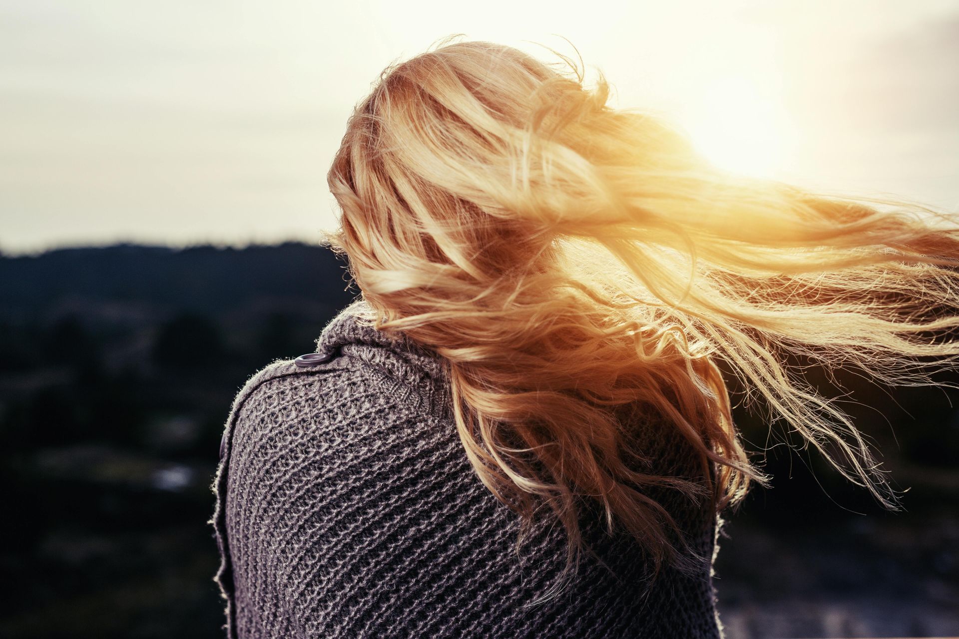 Woman with flowing blonde hair in sunlight, wrapped in a gray shawl, looking out at a blurred landscape.