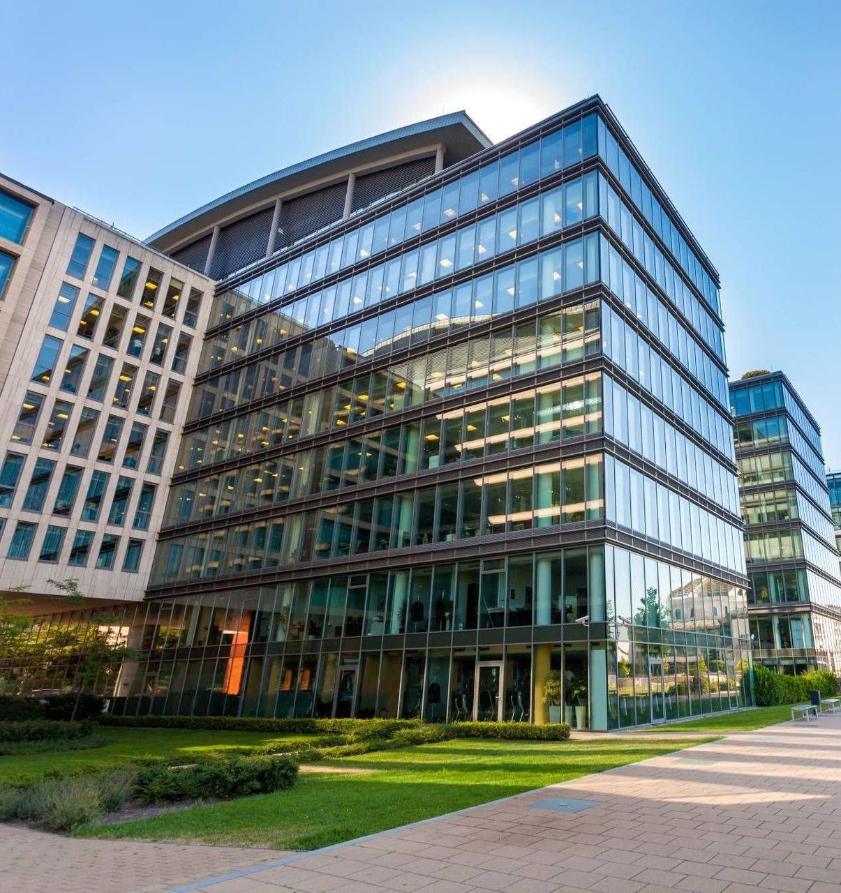 Modern glass-walled office buildings with a bright blue sky above, green lawn, and sun reflection.