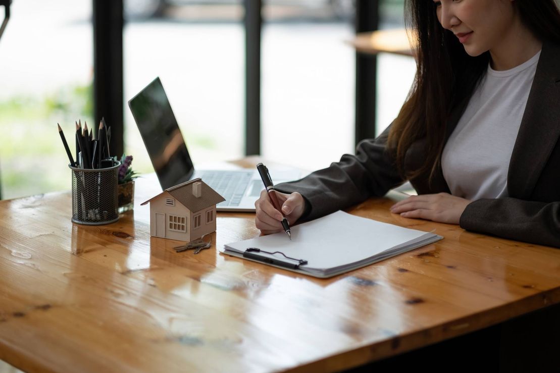 A woman is sitting at a wooden table writing in a notebook.