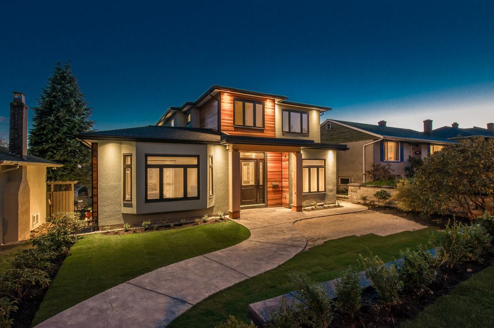 A modern two-story house at dusk, with illuminated facade, pathway, and lush landscaping.