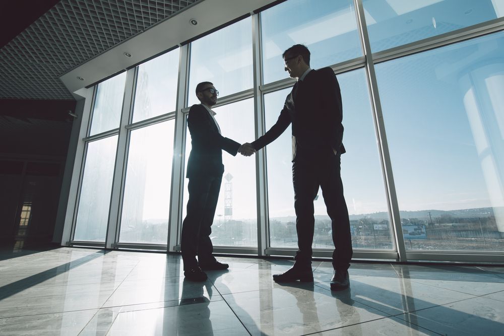Two businessmen are shaking hands in front of a large window.