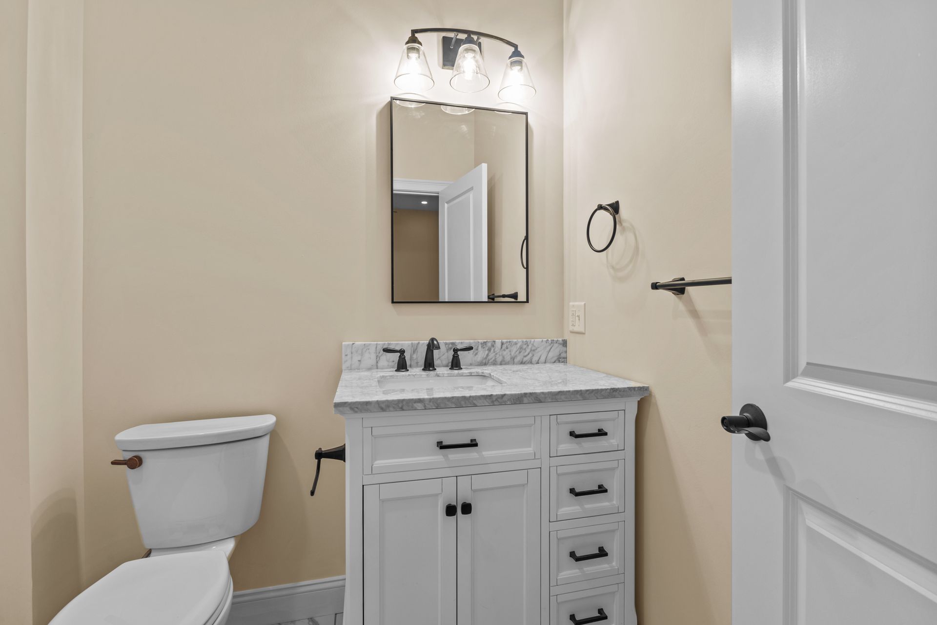 Small bathroom with white vanity, toilet, and light-colored walls. Black fixtures, mirror, and towel racks.