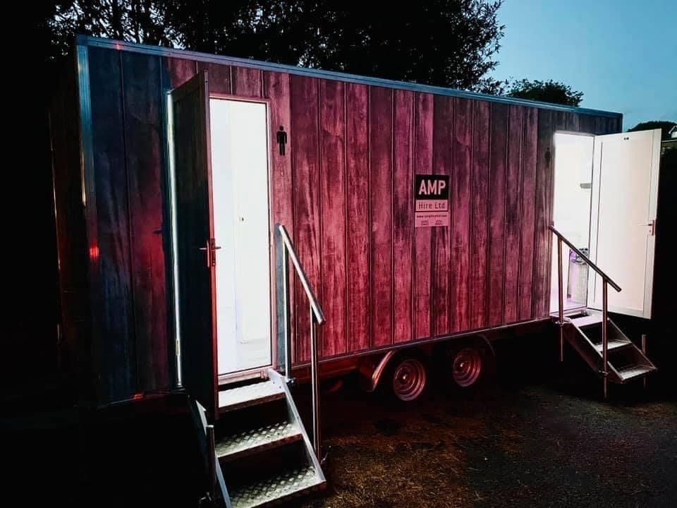 Portable restroom trailer with wood-grain facade. Two open doors with lit interiors and stairs. Dusk setting.