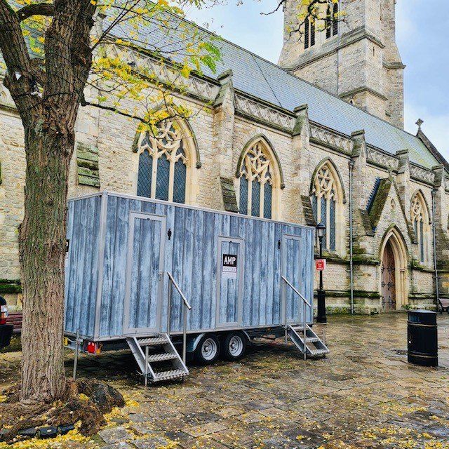 A mobile restroom trailer in front of a stone church. Gray wood exterior, metal steps, and a tree in view.