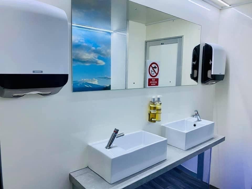 Modern restroom with white sinks, mirror, soap dispenser, paper towel dispenser.