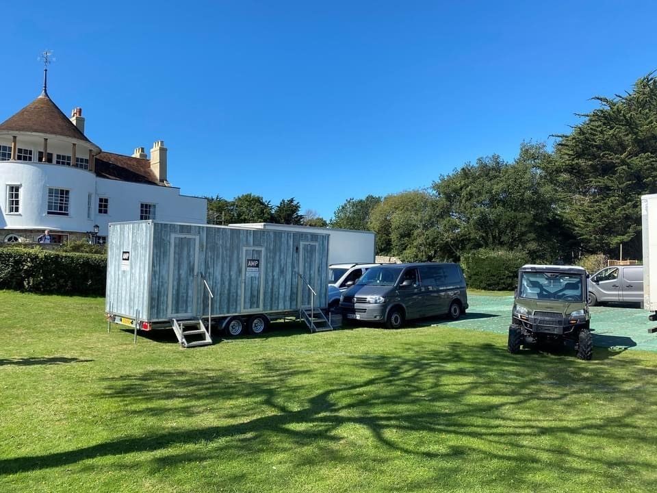 Portable toilet trailer and vehicles on a grassy lawn near a white building with a turret on a sunny day.