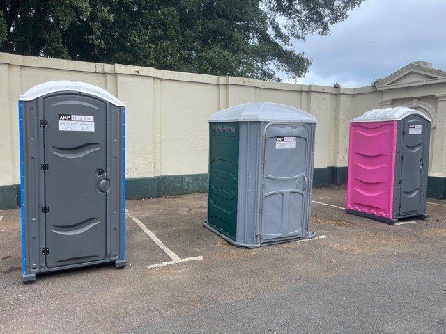 Three portable toilets in a parking area against a white wall; one gray with blue trim, one green, one pink.