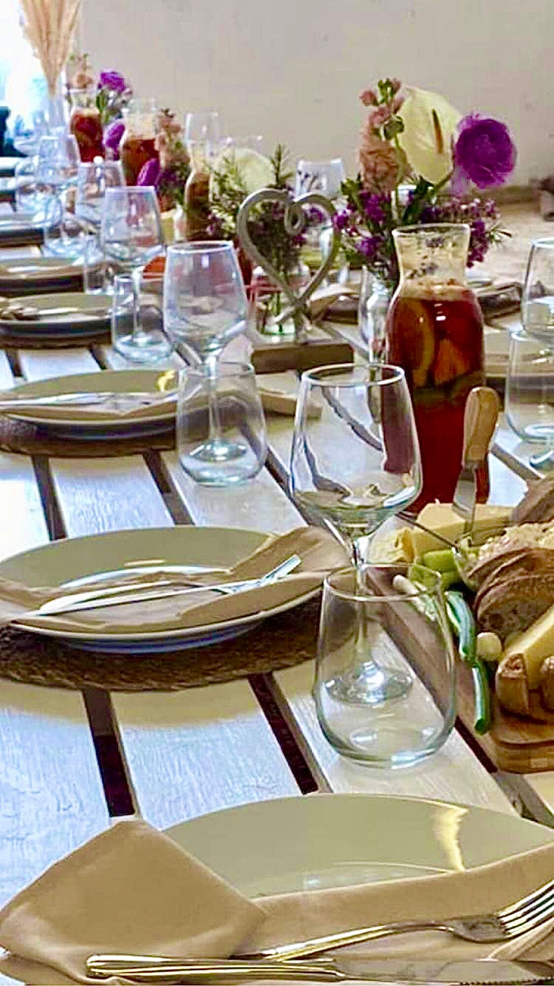 Long table set with food, drinks, and floral arrangements for a gathering.