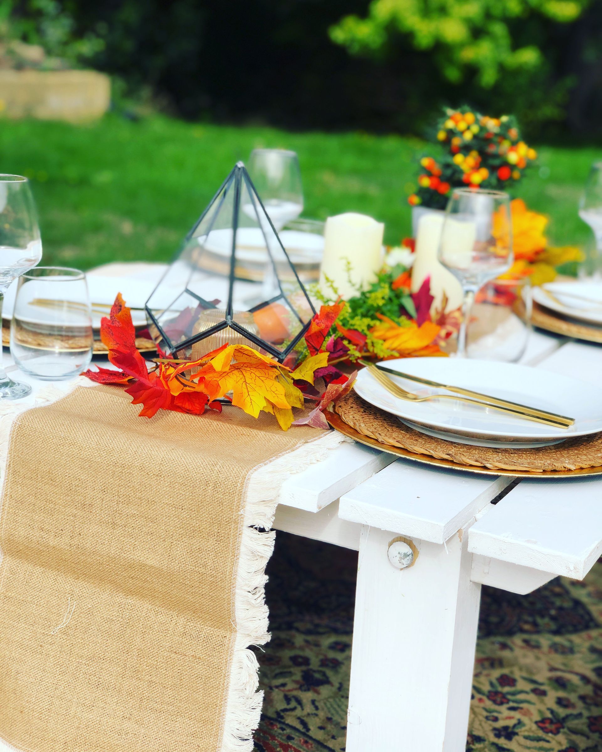 Fall picnic table setting with autumn leaves, candles, and geometric glass decor on white wood.