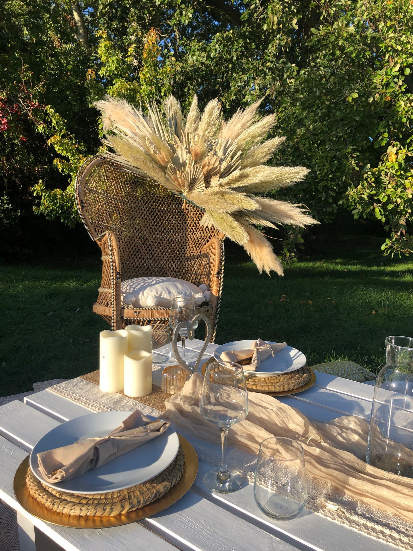 Bohemian outdoor dining setting. A wicker chair with pampas grass decor, table with plates and candles.