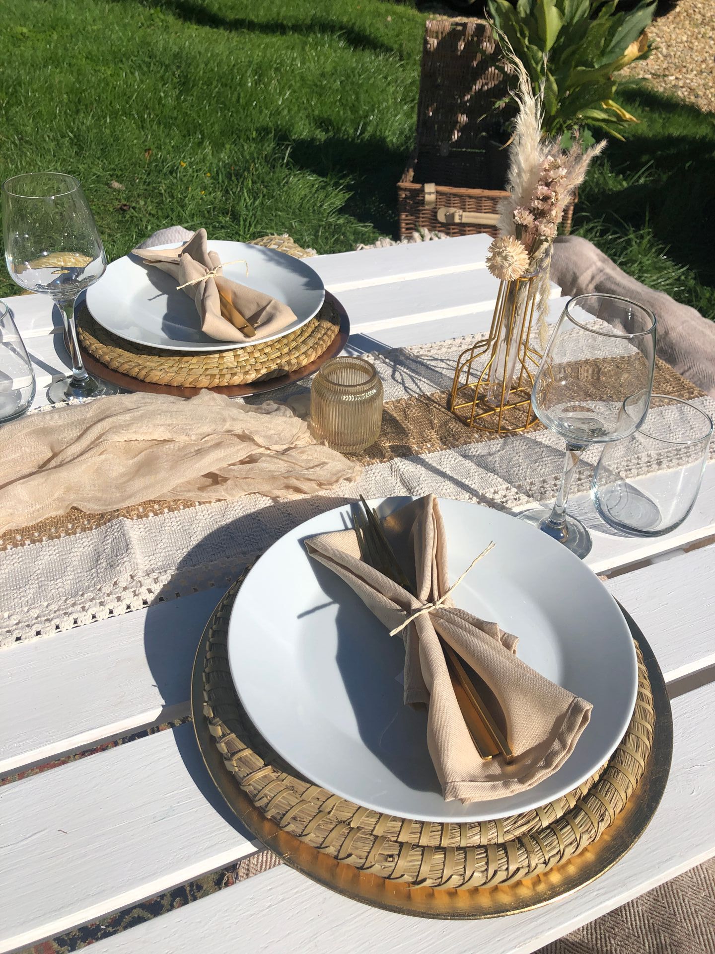 Outdoor picnic setting with white plates, woven chargers, and folded napkins on a white wooden table.
