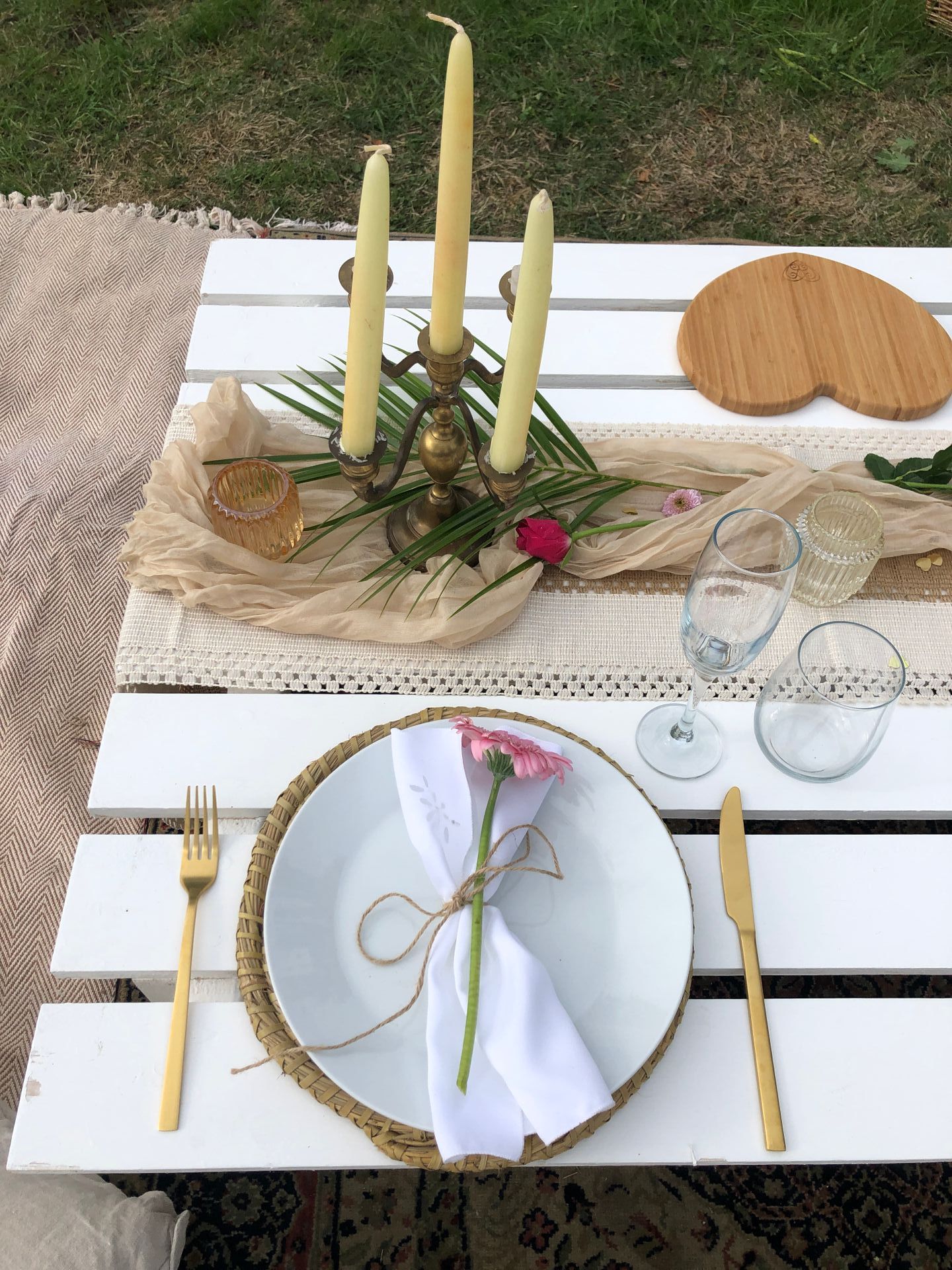 Picnic table set for a meal: white plates, gold utensils, candles, flowers, glasses, and a woven placemat.
