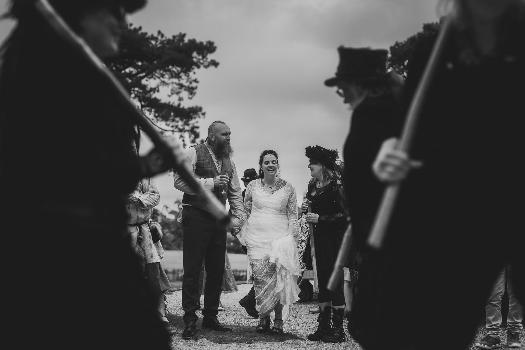 Bride and groom walk between costumed figures holding staves, smiles, outdoor setting, black and white.