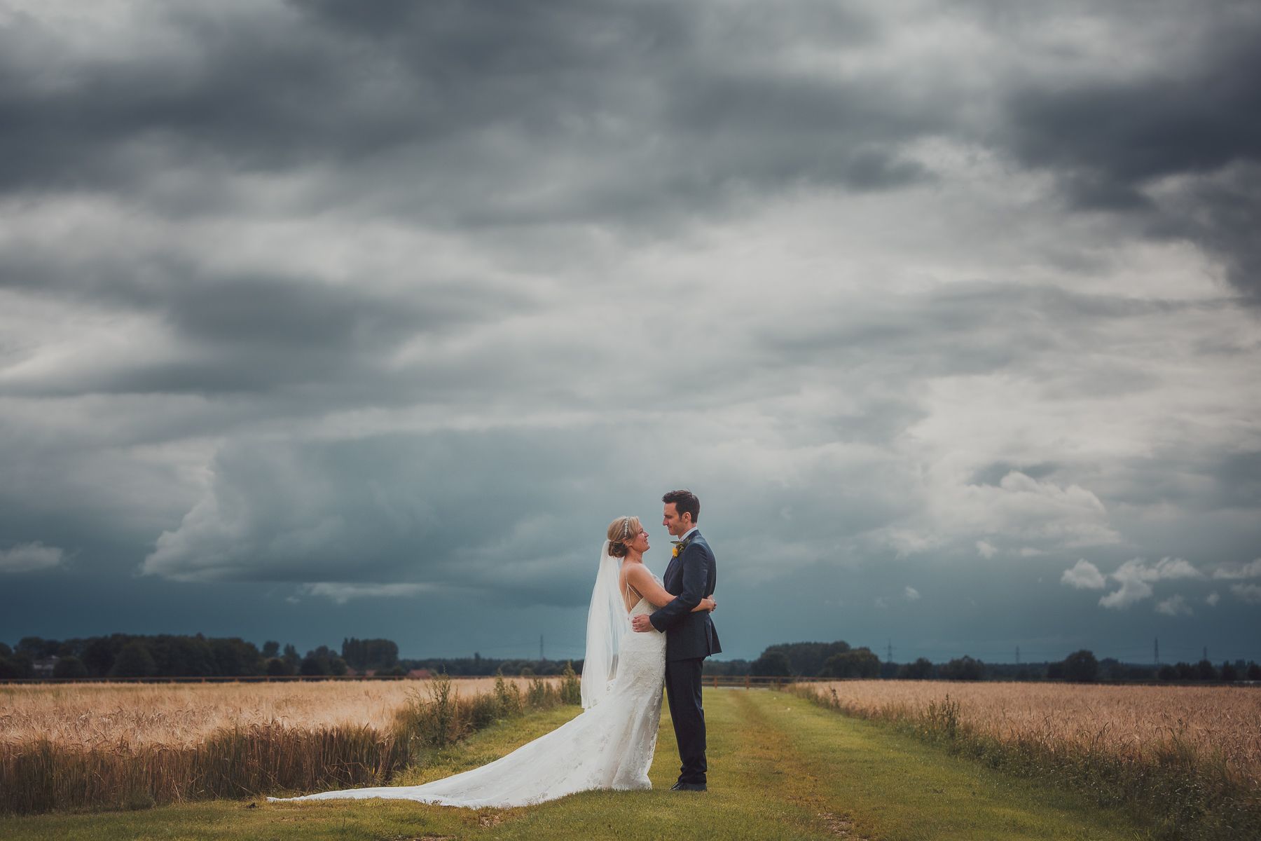 Bride and groom embrace on a path through a field, dramatic dark sky overhead.