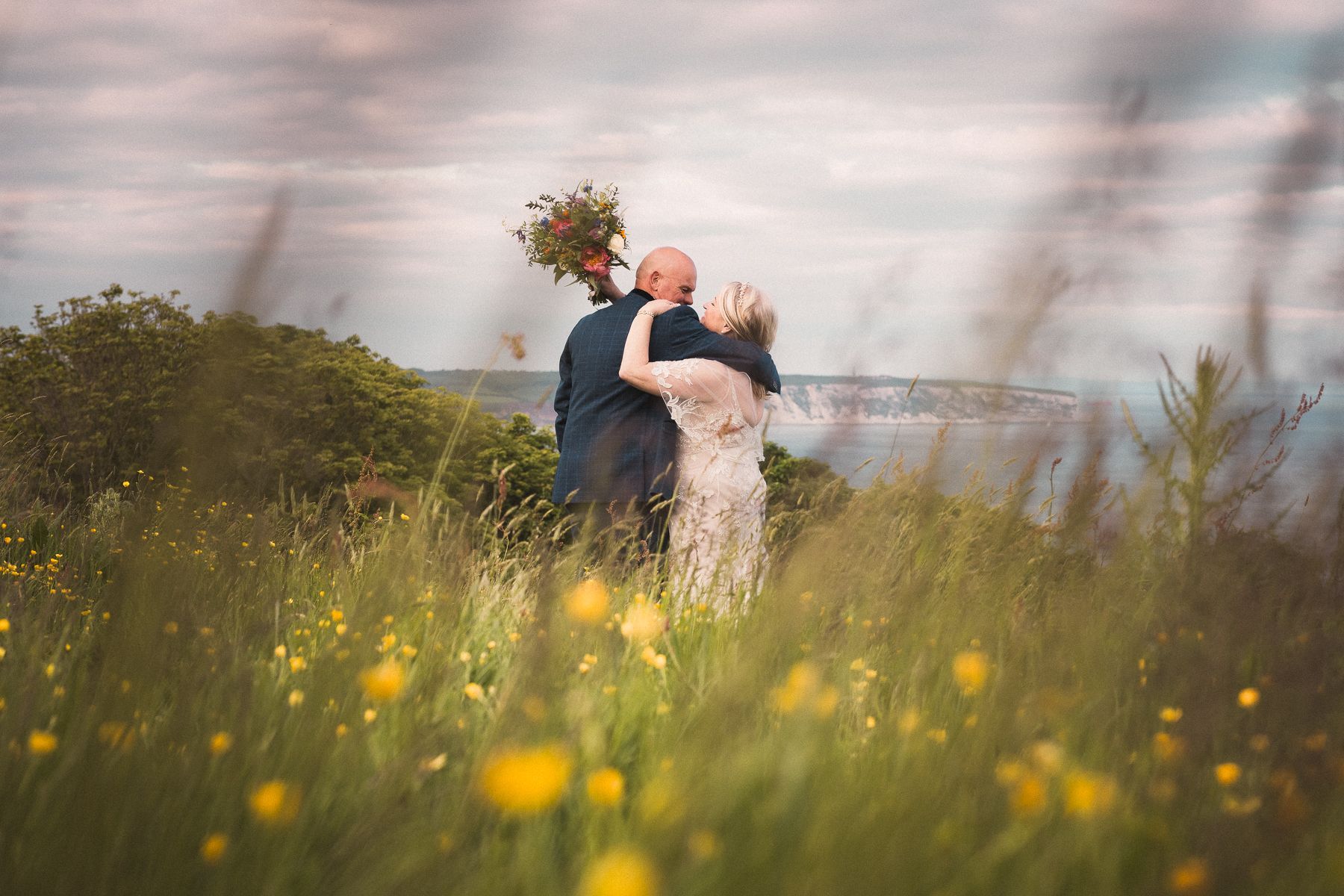 Couple embracing, holding bouquet, in a field of yellow flowers, overlooking water. Overcast sky.