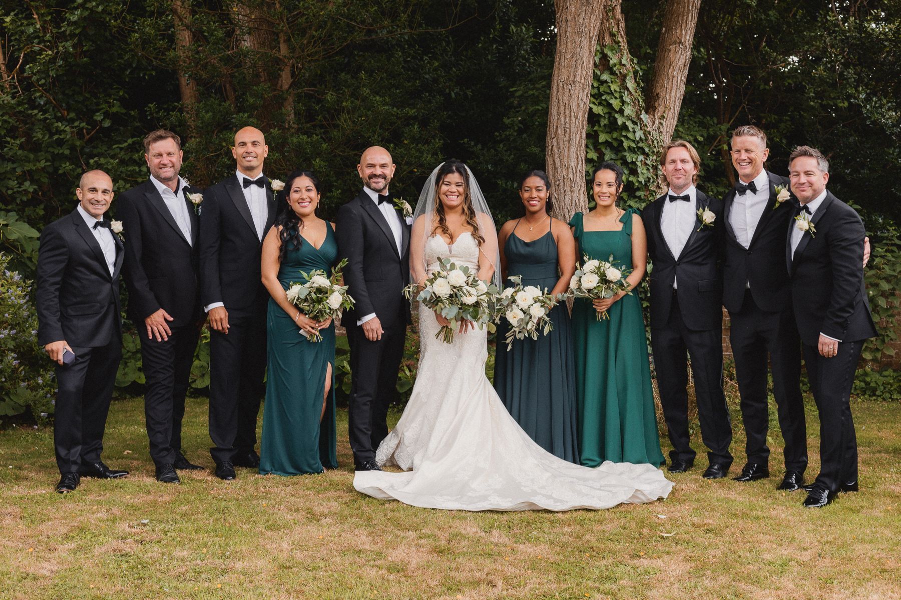Wedding party posing outdoors; bride in white gown, bridesmaids in green dresses, groomsmen in black suits.