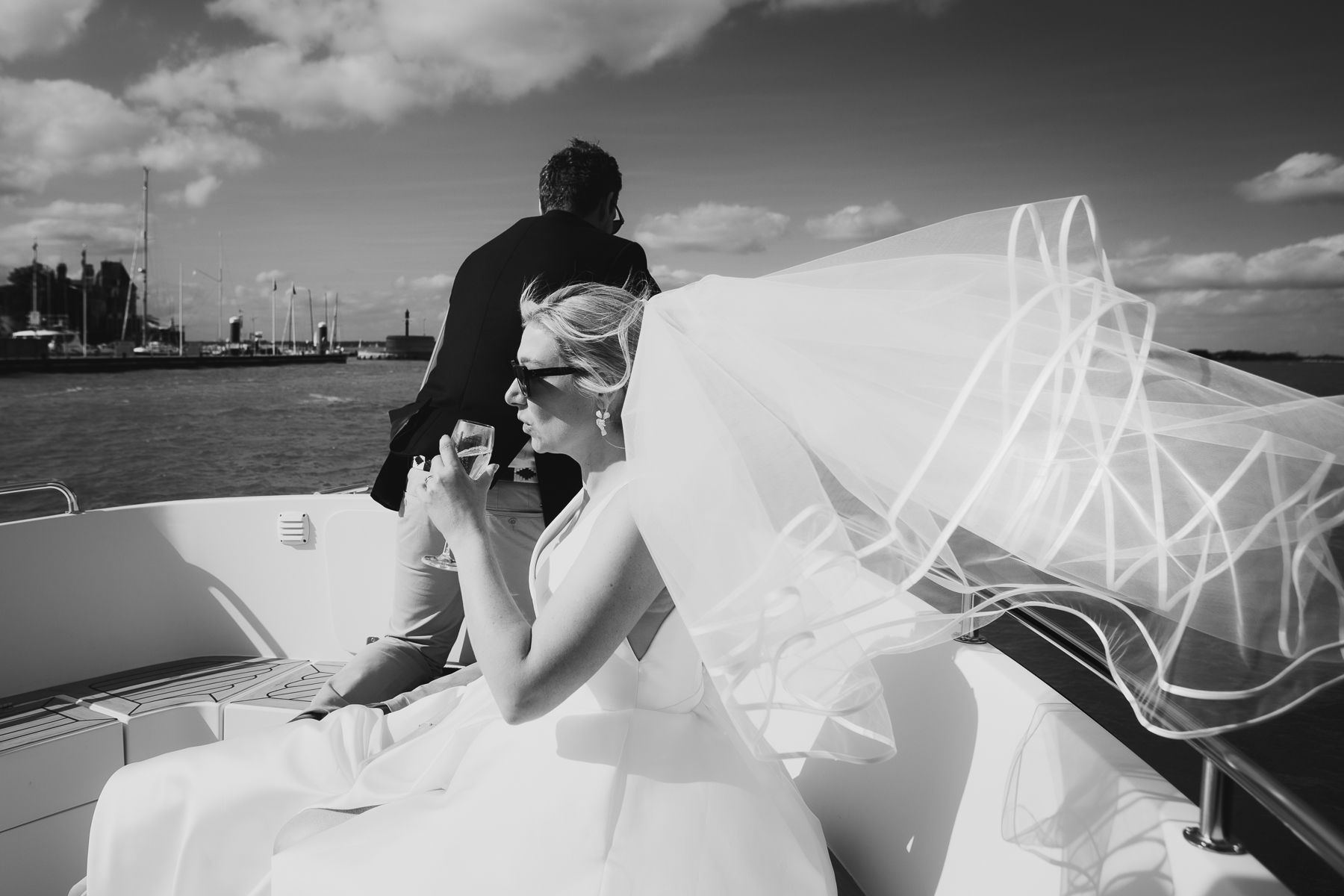 Bride in wedding dress and veil on boat, holding drink, with groom in background.