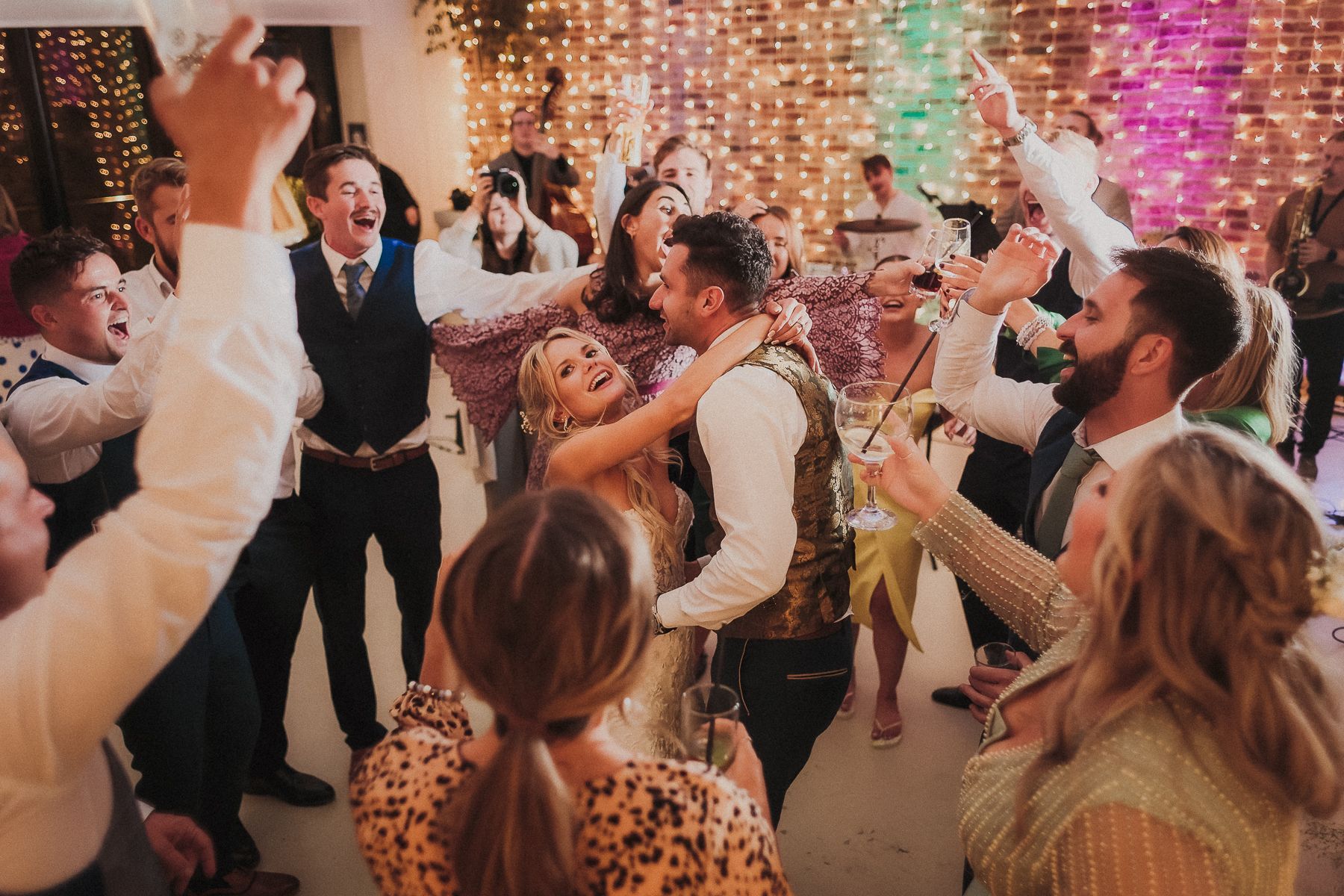 People celebrating at a wedding reception, couple dancing surrounded by guests raising drinks, string lights in background.