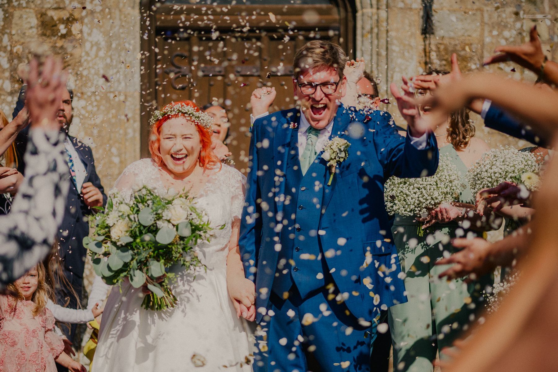 Bride and groom exiting a building, covered in confetti, joyfully smiling.