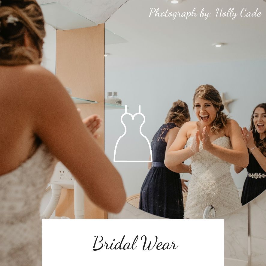 Bride in a wedding dress looking in a mirror with friends, clapping.