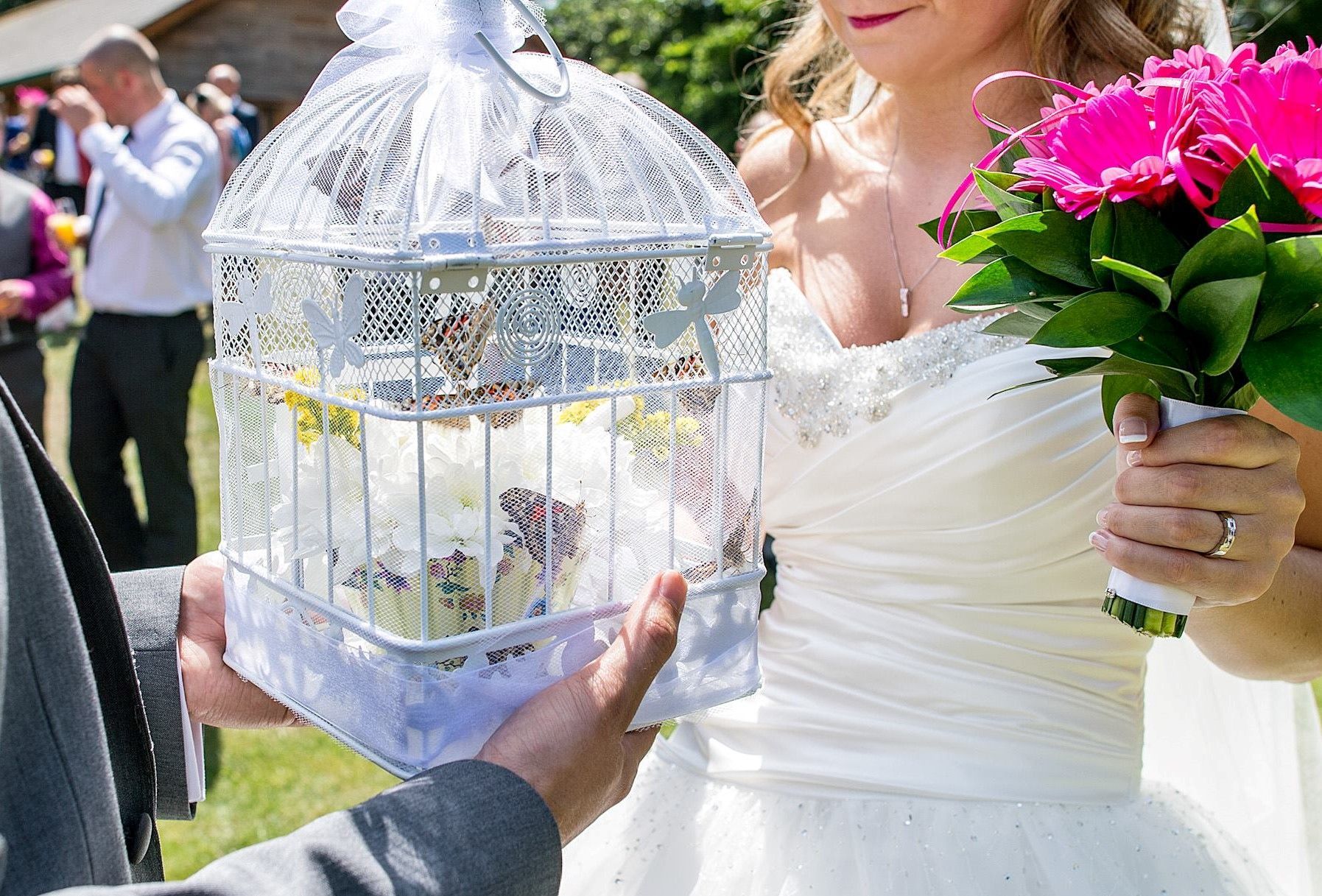 Bride holding a birdcage filled with cupcakes and a pink flower bouquet at an outdoor wedding.