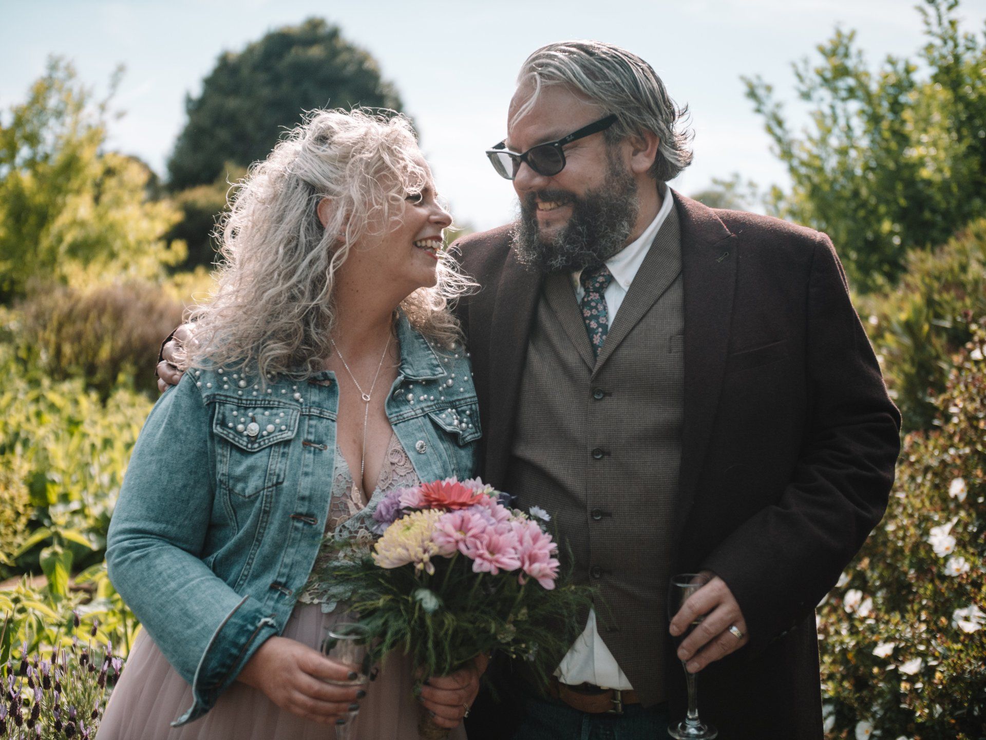 Couple, smiling and embracing outdoors. Woman in pink dress, denim jacket, holding flowers. Man in suit jacket, sunglasses.