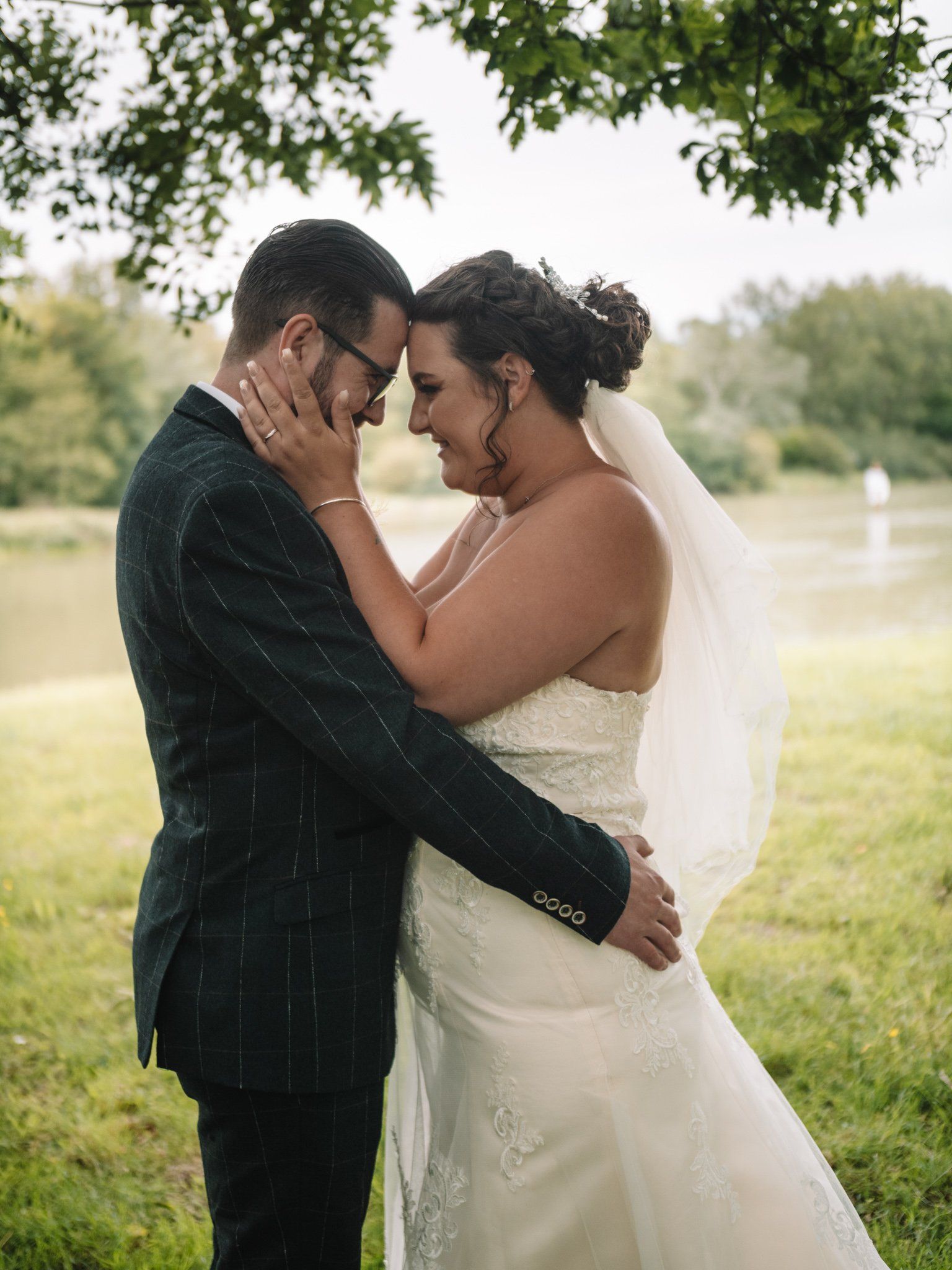 Wedding couple embracing outdoors; man in suit, woman in white dress and veil, touching faces near a lake.