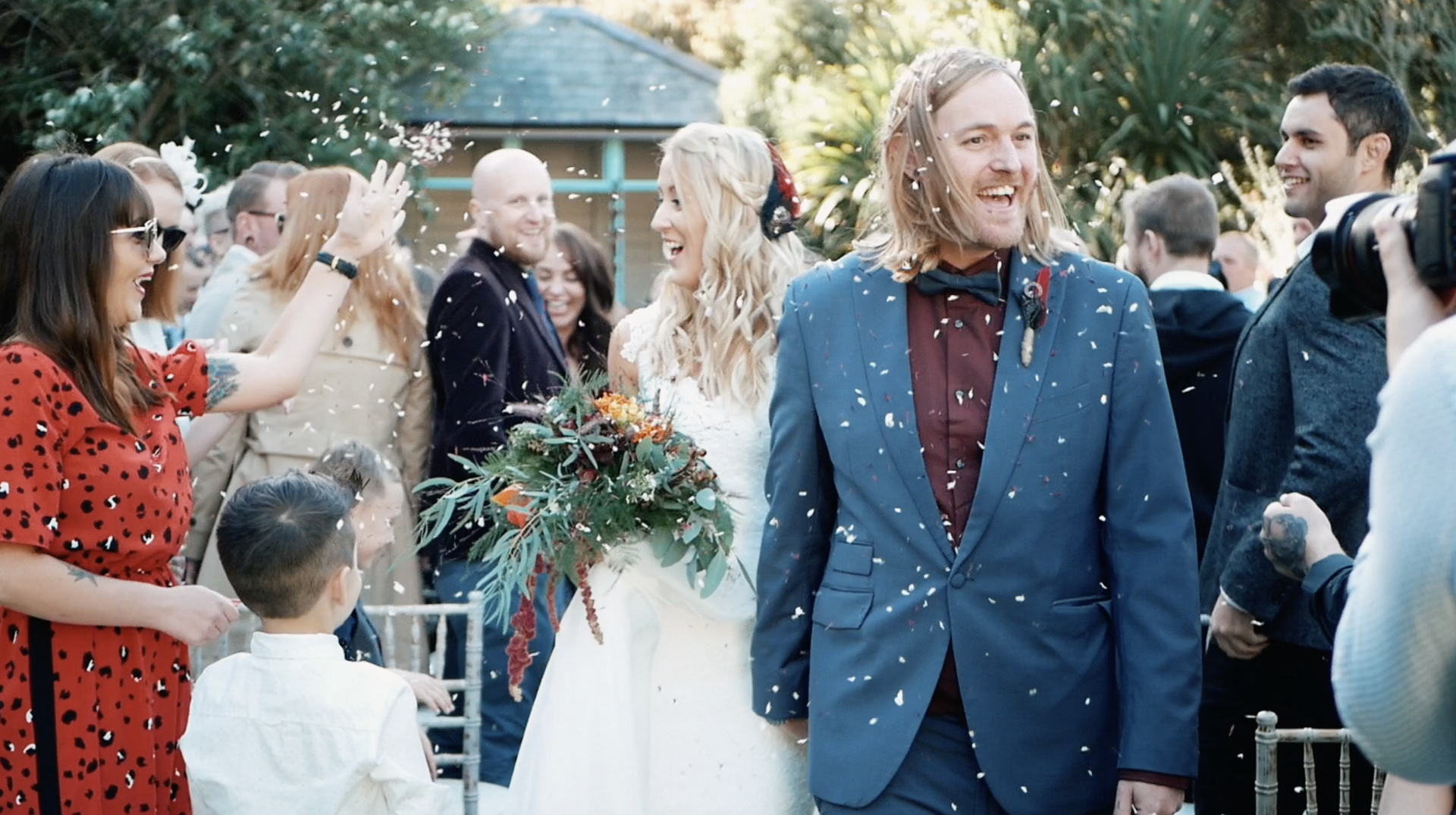 Newlyweds walk through confetti, smiling, with guests in outdoor wedding setting.