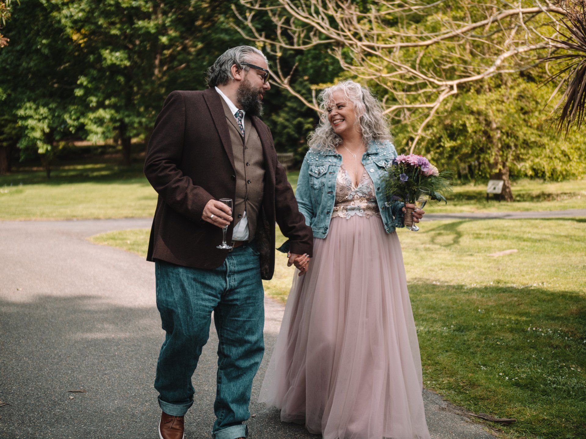 Couple holding hands, smiling while walking on a path in a park. One in wedding dress, the other in a suit jacket and jeans.