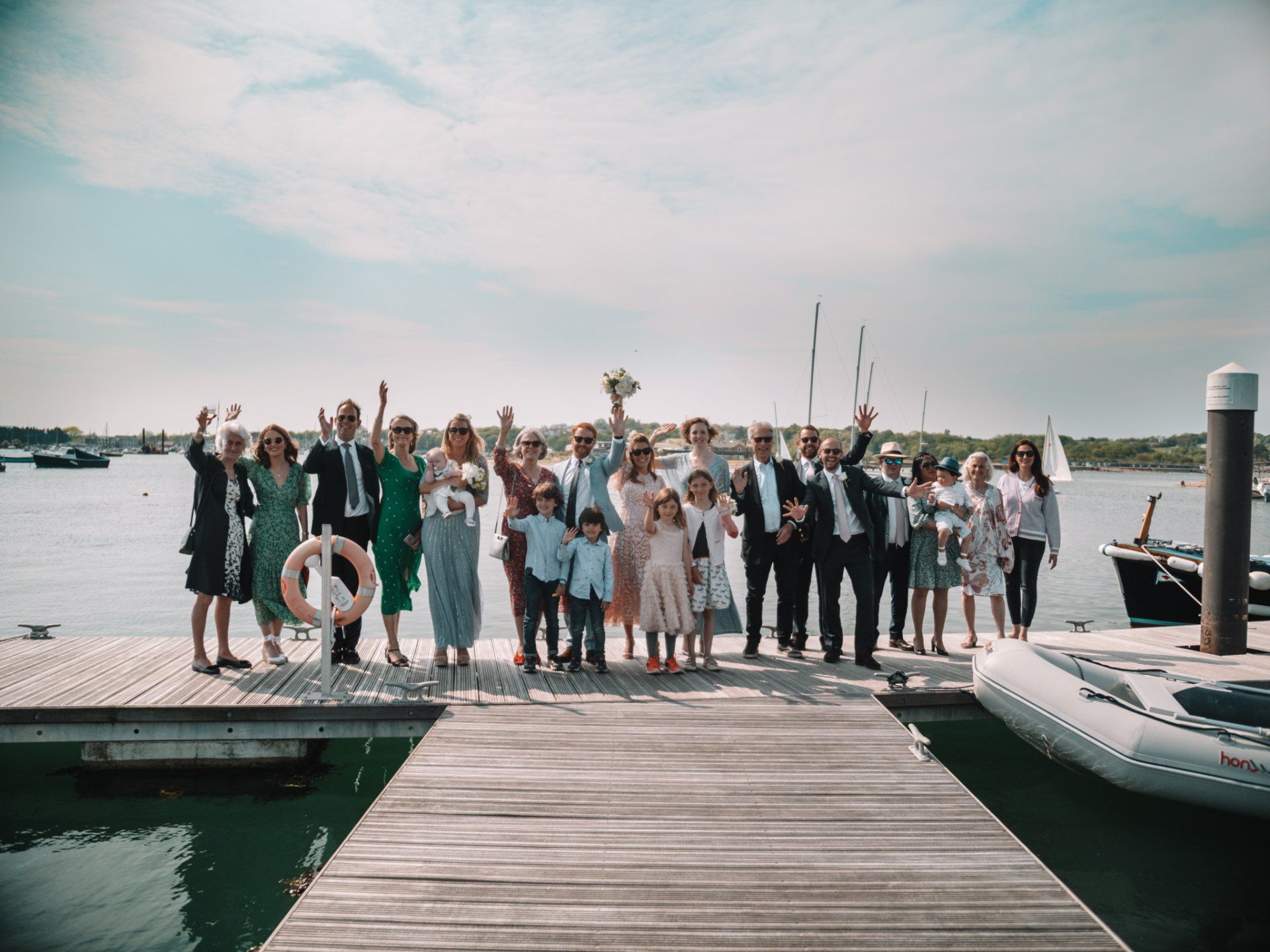 Wedding group waves on a wooden dock with boats, sky, and water.