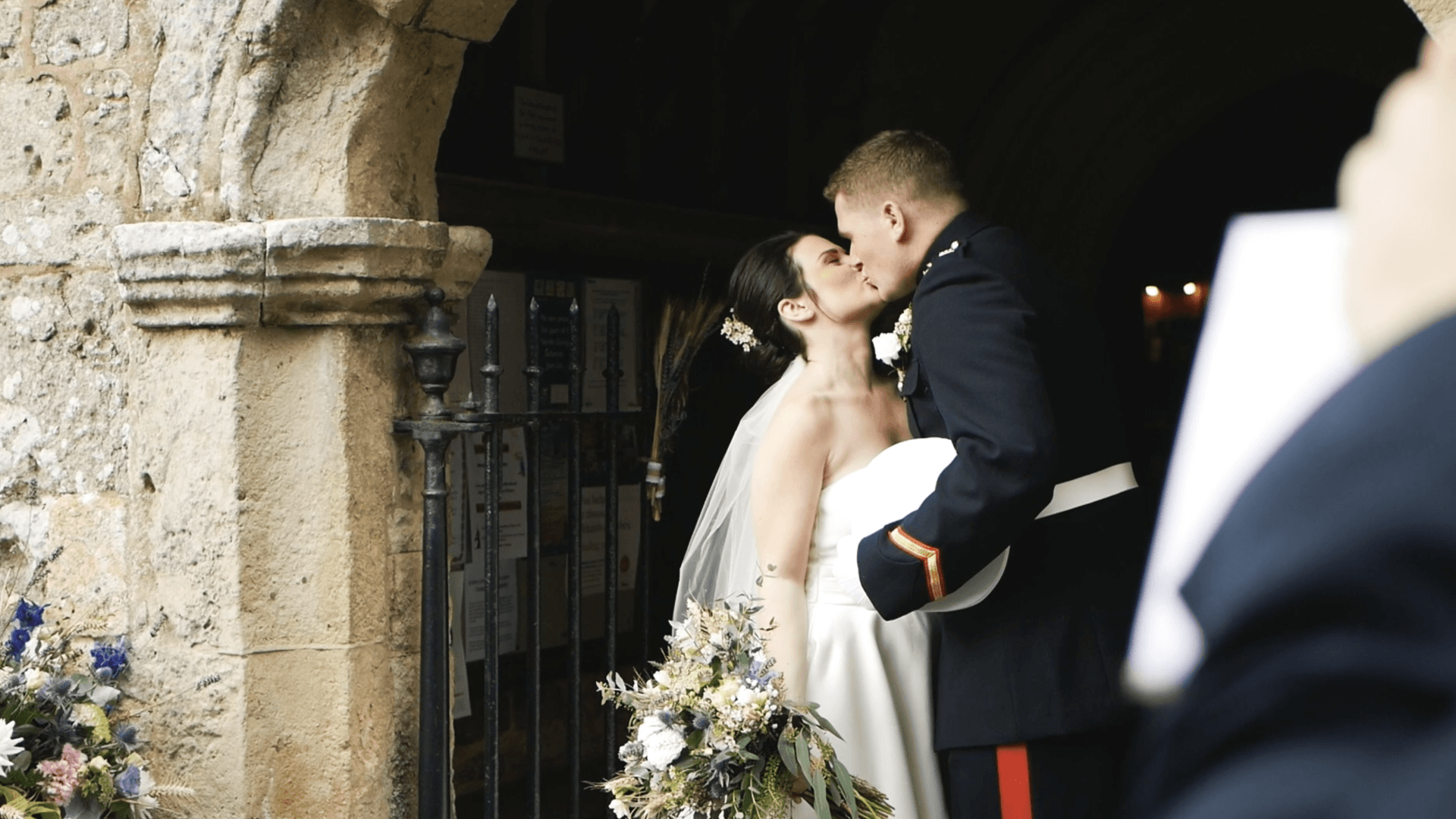 Newlyweds kiss in a church doorway. The groom wears a military uniform, and the bride wears a veil and holds a bouquet.