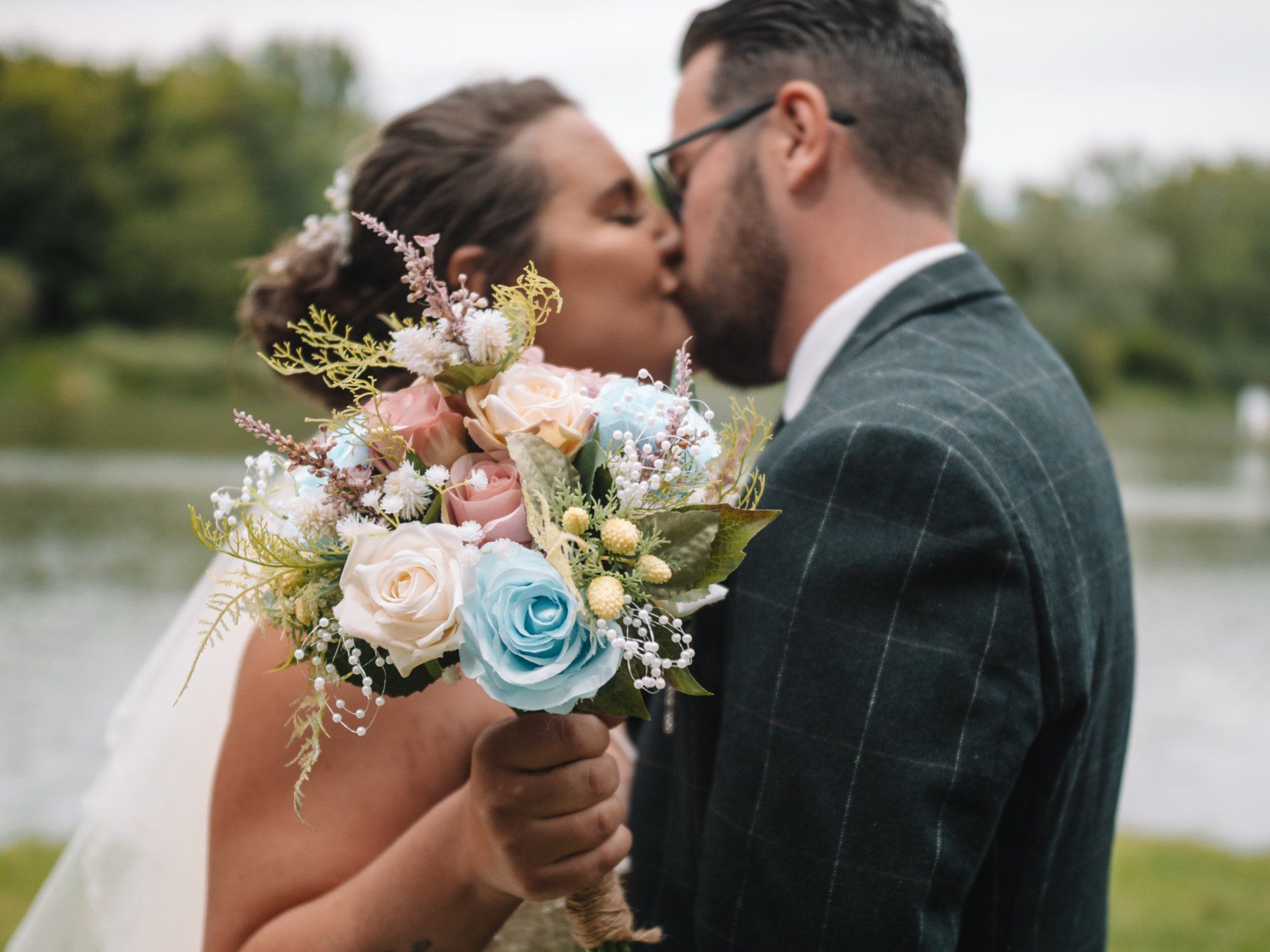 Bride and groom kissing, holding bouquet with blue and peach flowers, near a lake.