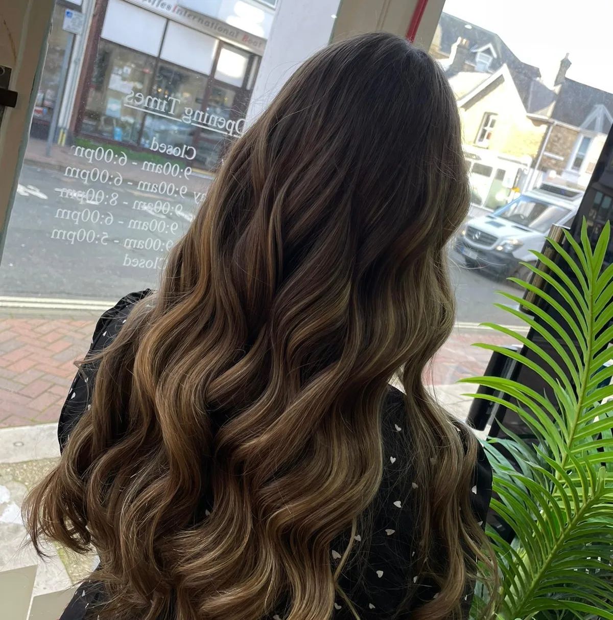 Woman with long, wavy brown hair, looking away, in a salon, sunlight illuminating the hair.