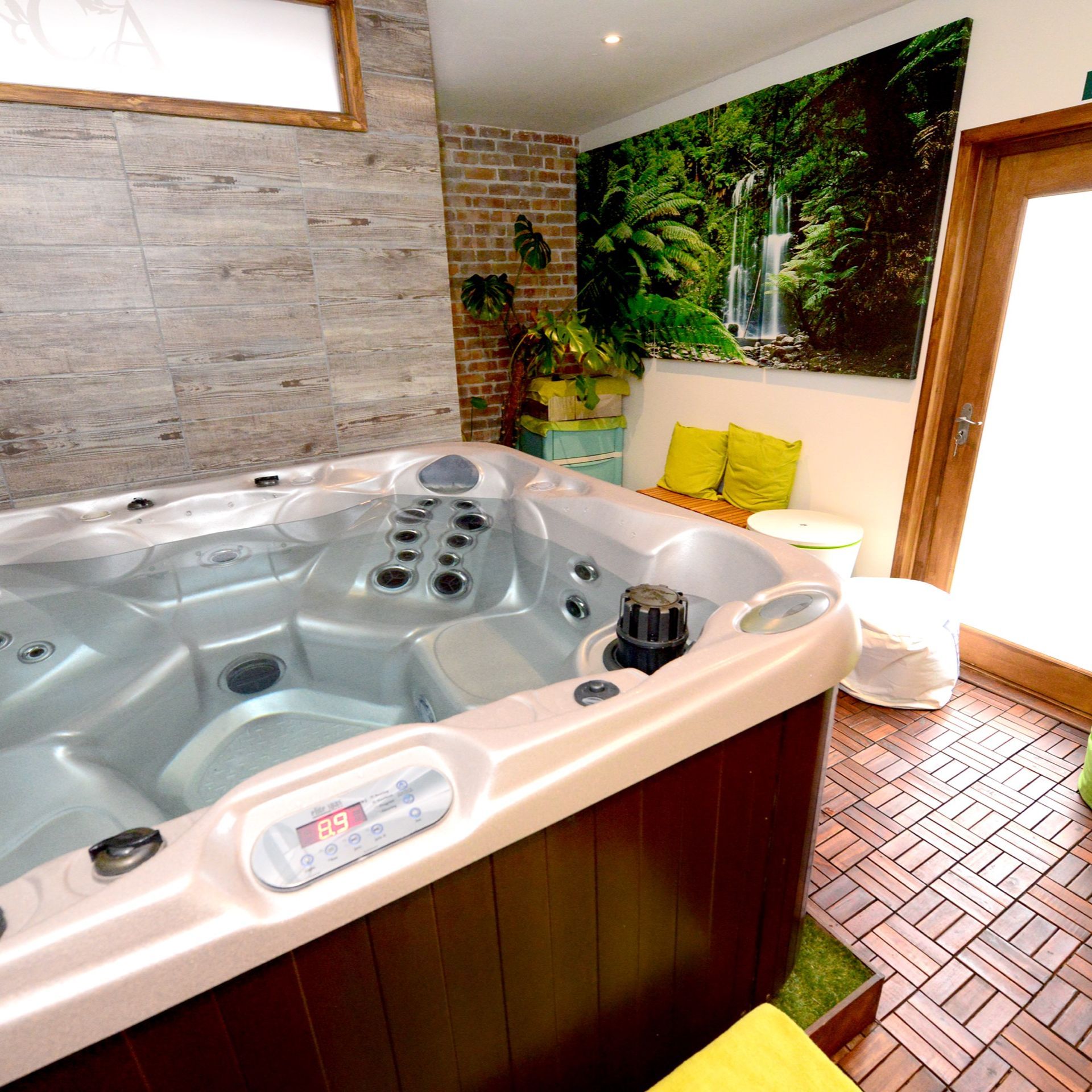 Hot tub in spa setting, with brick wall, waterfall mural, and wooden flooring.