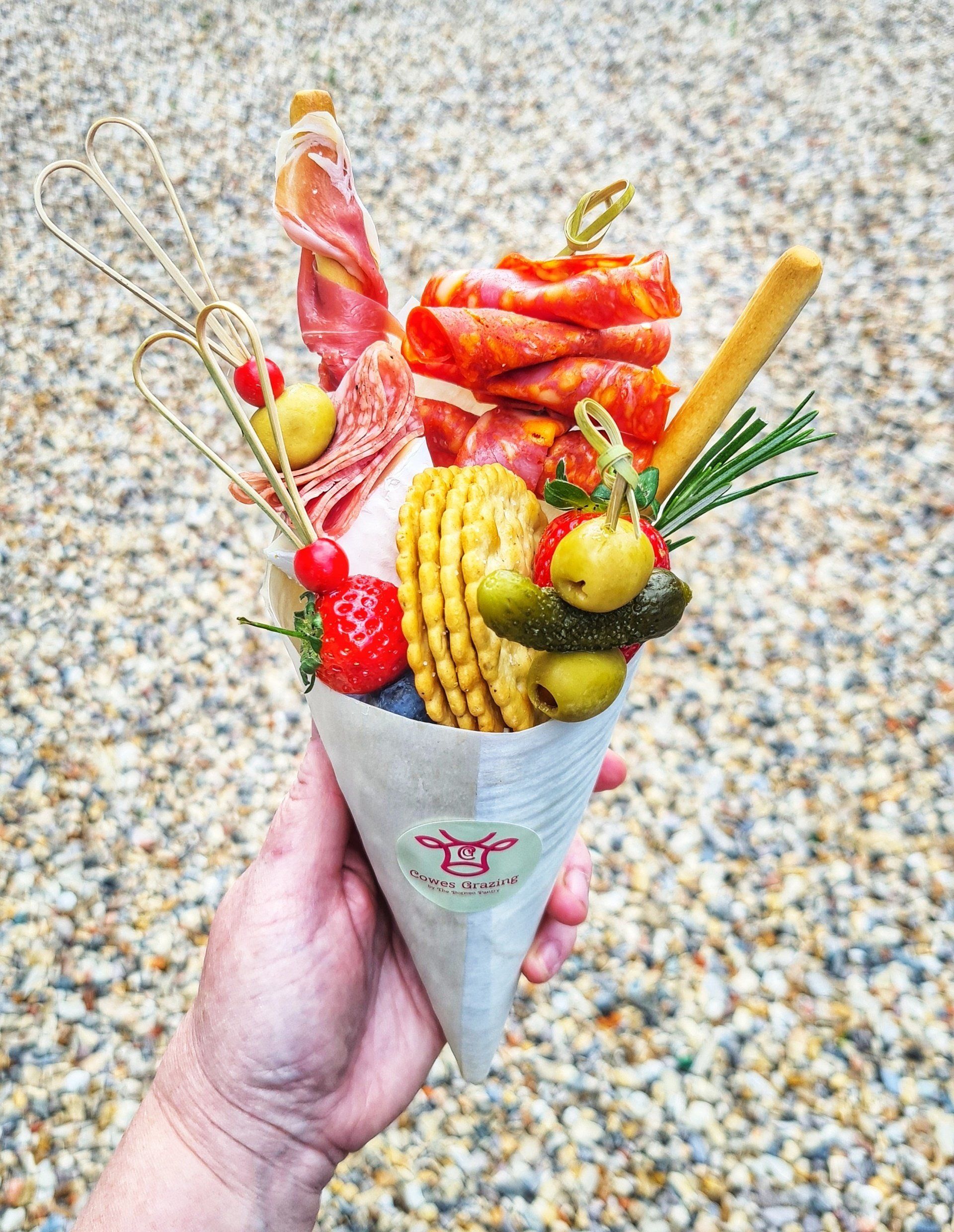 Person holding a charcuterie cone filled with meats, crackers, olives, and garnishes.