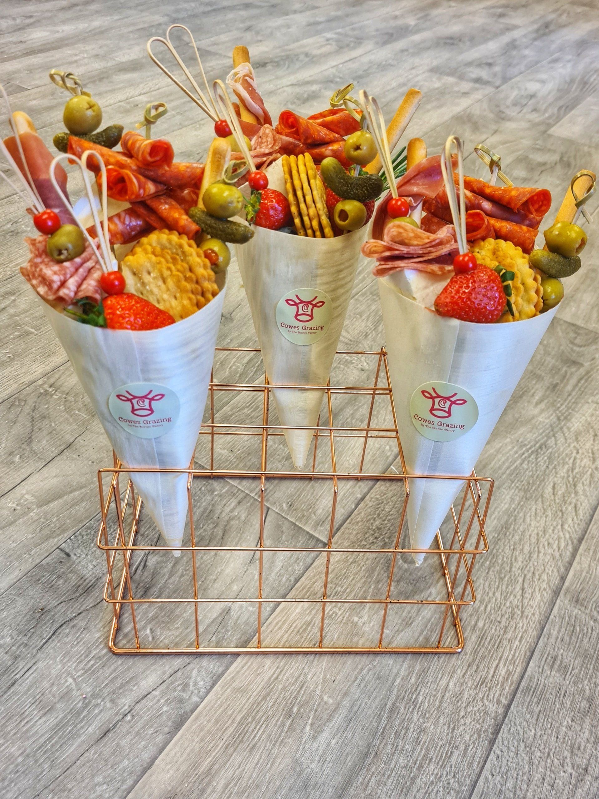 Charcuterie cones in a rose gold wire holder on a grey surface. Each cone contains meats, crackers, and olives.