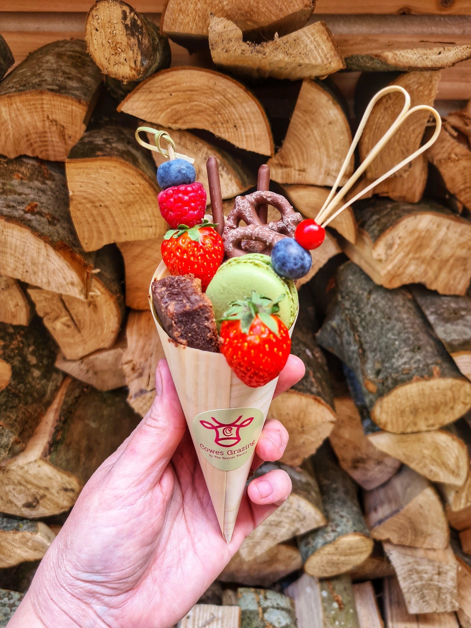 Hand holding a decorated ice cream cone with berries, chocolate, and pretzels, against a woodpile backdrop.