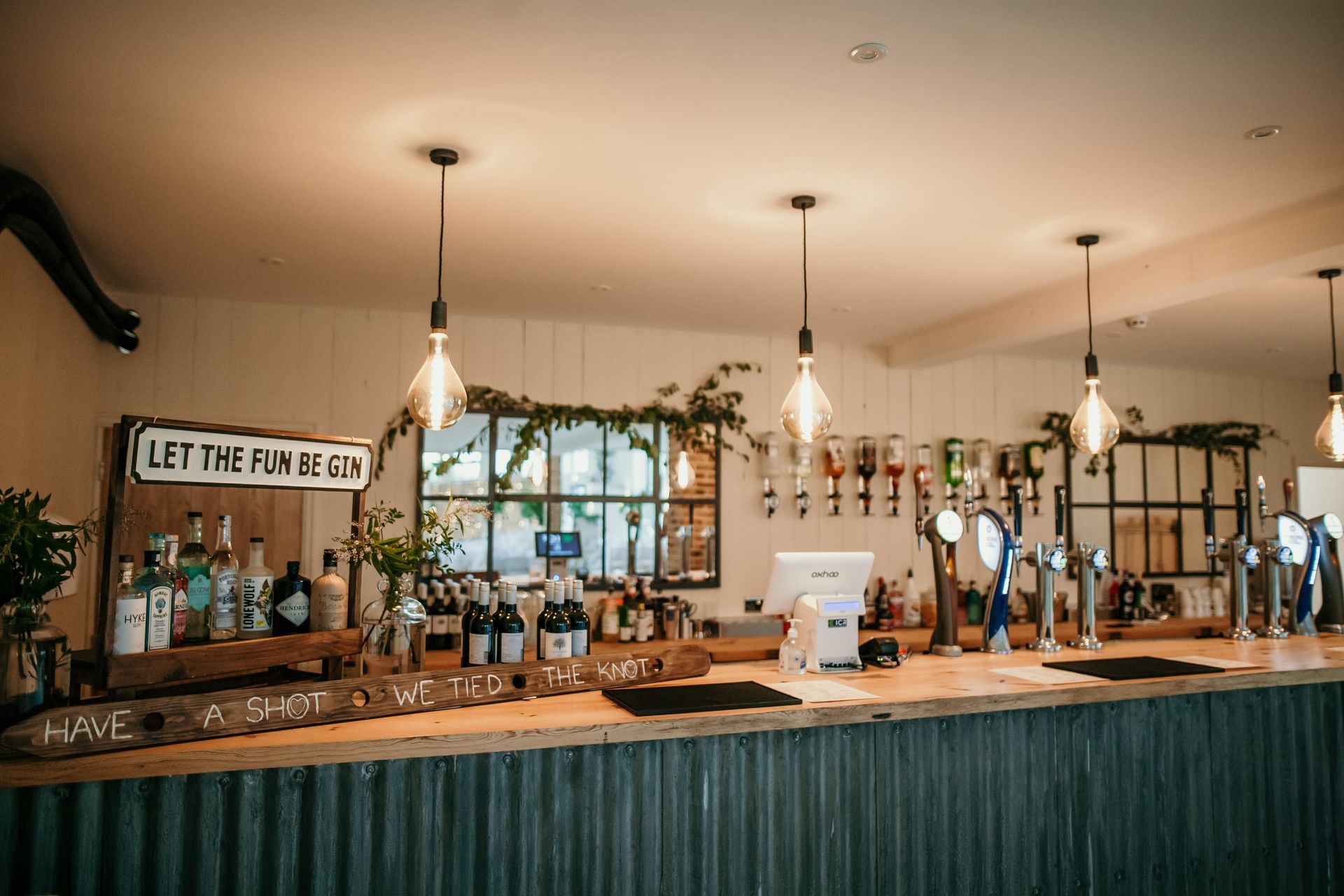 Bar with drinks, bottles, and hanging lights; rustic decor, windows in background.