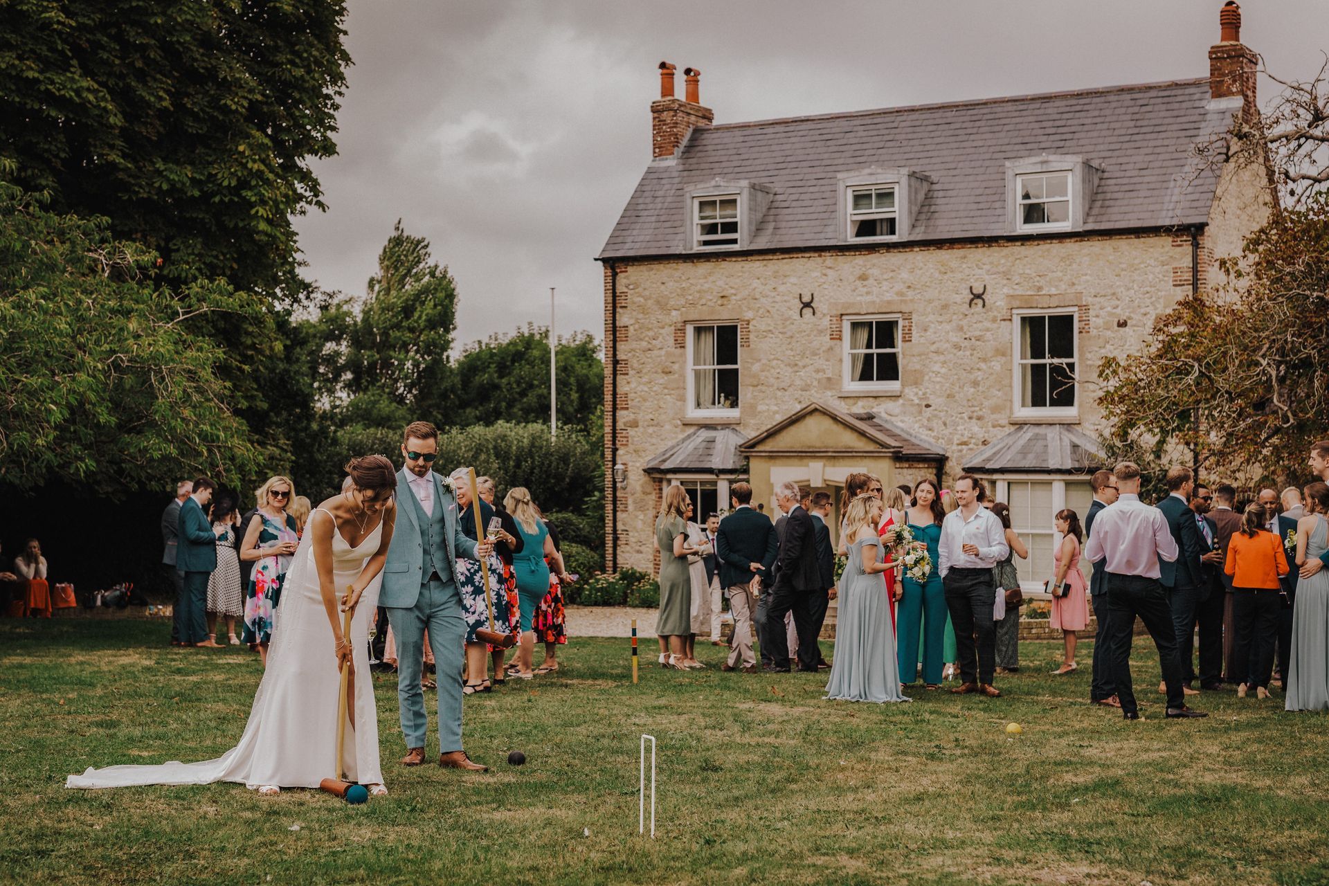 Bride and groom playing croquet on a lawn, guests watching near a large house. Cloudy sky.