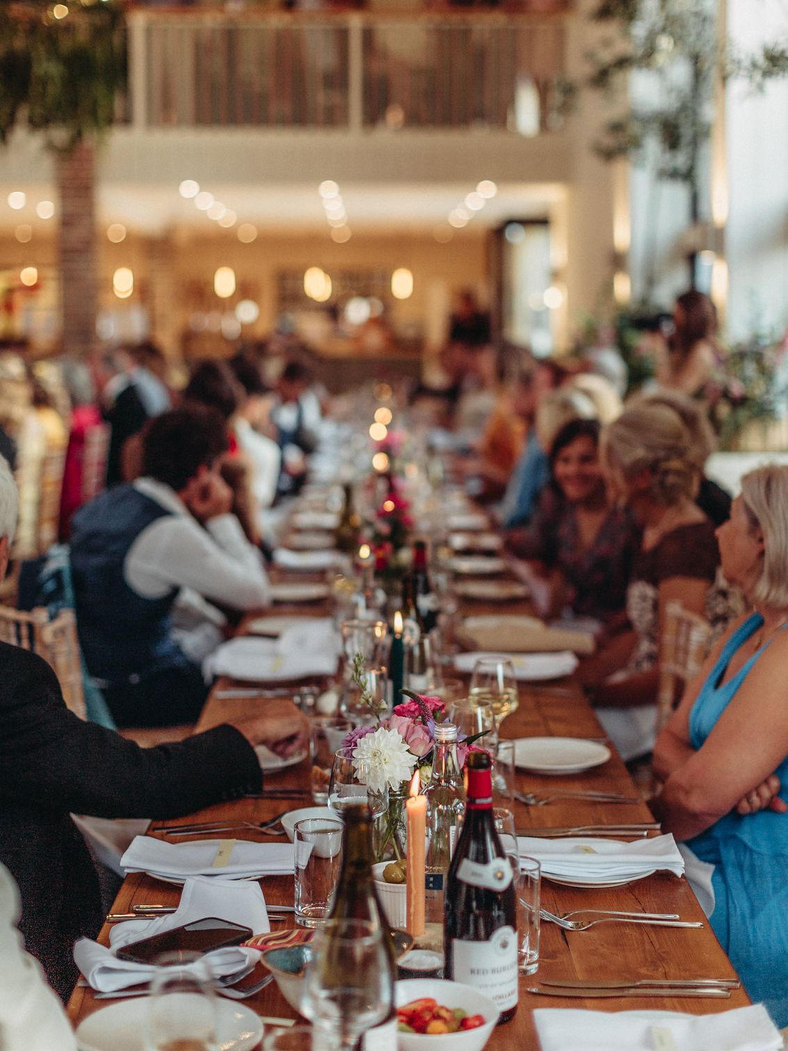 Long table set for a celebration, guests seated, candles, flowers, wine bottles, bright interior.