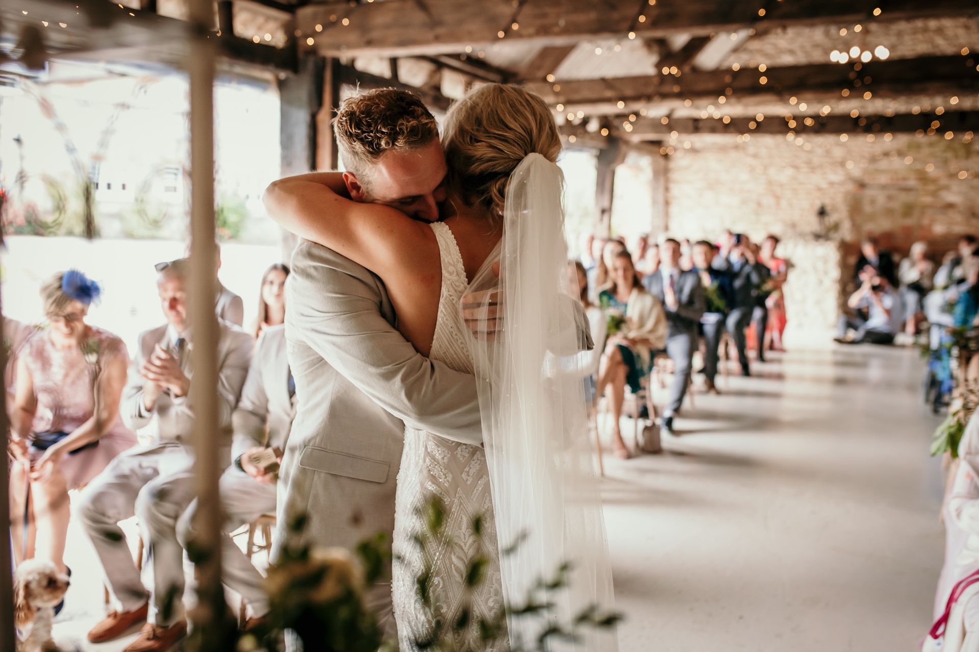 Bride and groom embrace, kissing, in a barn during a wedding ceremony. Guests watch.